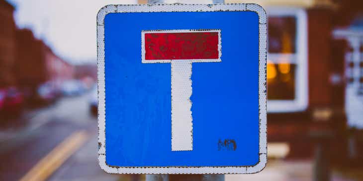 Ein verrostetes, blau-rot-weißes Straßenschild, hängt an einem Pfeiler und erinnert an den Buchstaben „T“.