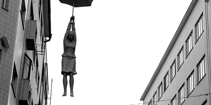 Awangardowa rzeźba kobiety zwisającej z parasolki zawieszonej pomiędzy budynkami.