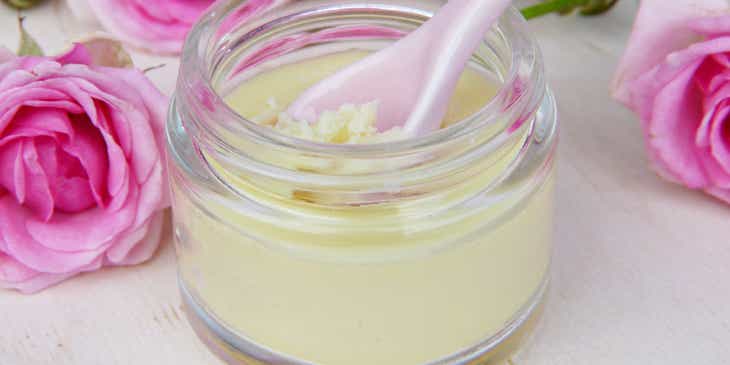 Sebotol body butter kecil dengan sendok merah muda dikelilingi mawar merah muda di bisnis body butter.