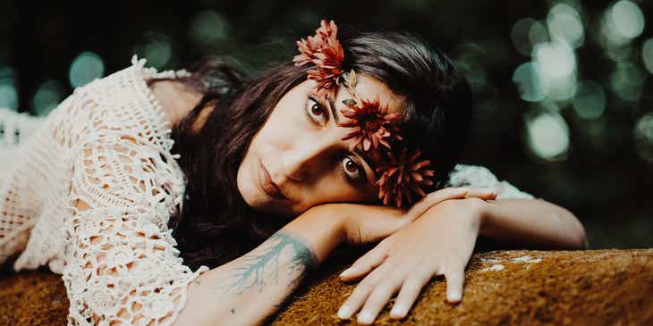 Uma mulher com estilo boho com o pulso tatuado e usando um top de crochê e uma coroa de flores deitada sobre uma pedra.