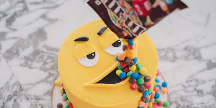 A custom cake of a yellow M&M with a bag of M&Ms being poured onto it.