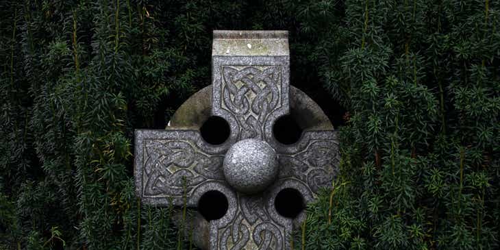 Ein keltisches Kreuz aus grauem Stein mit Ornamenten vor grünen Zweigen.