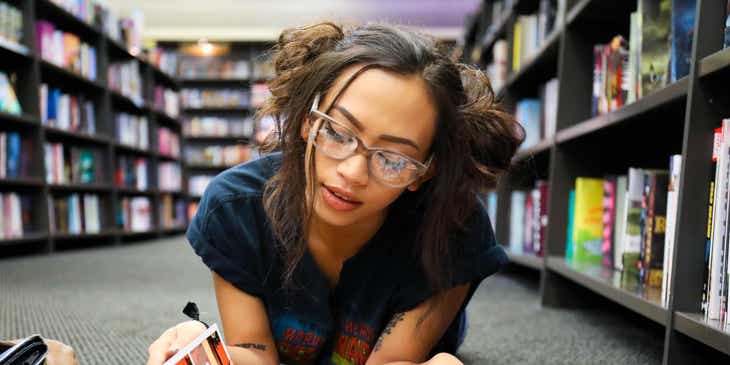 A geeky woman in glasses reading a comic on the floor of a bookshop.