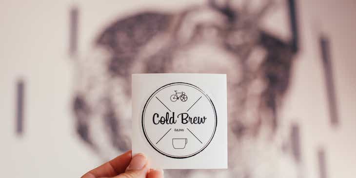 Osoba trzymająca naklejkę z logo dla kawiarni, w które zainwestowała po analizie kosztów.
