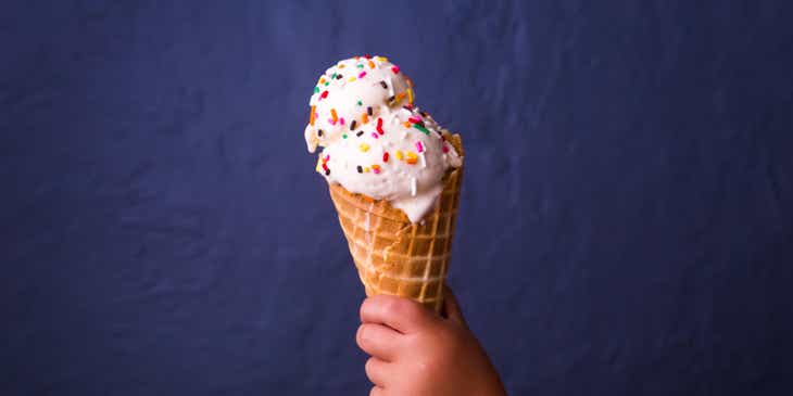 Tangan seorang balita memegang sebuah cone es krim dengan dua scoop es krim dan topping.
