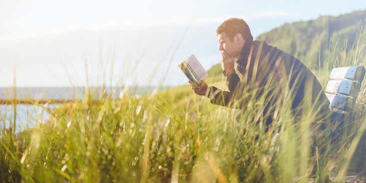 Eine intelligente Person sitzt neben dem Meer in einem überwachsenen Feld auf einer Bank und liest ein Buch.