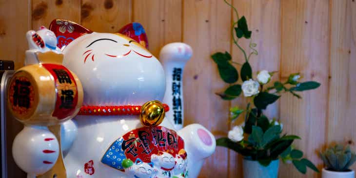 Ein chinesischer Glücksbringer in Form einer Katze steht auf einem Tisch neben einer Topfpflanze.