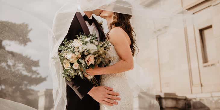 Uno sposo e una sposa che si baciano sotto un velo il giorno del loro matrimonio.