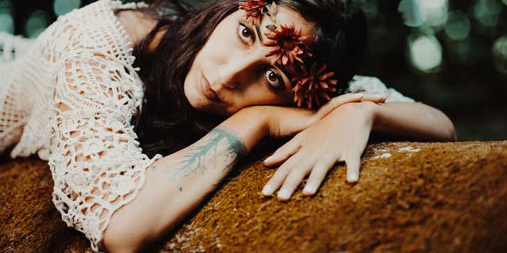 Een boho-vrouw met getatoeëerde pols in gehaakt topje en bloemenkroon, liggend op een rots.