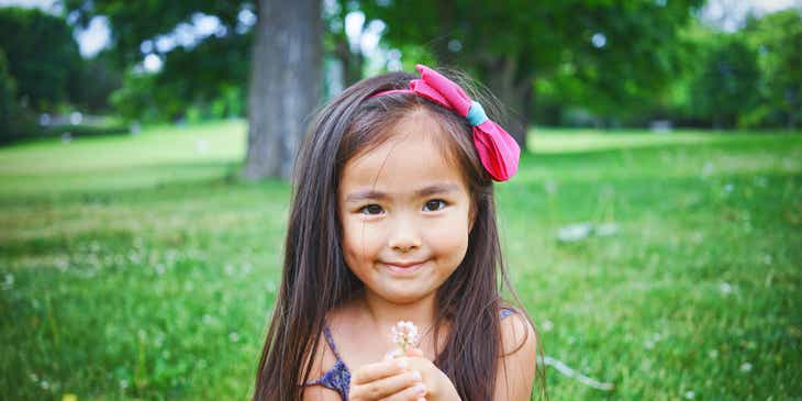Ein junges Mädchen mit einer pinken Haarschleife hält in einem Feld eine Pusteblume in beiden Händen.