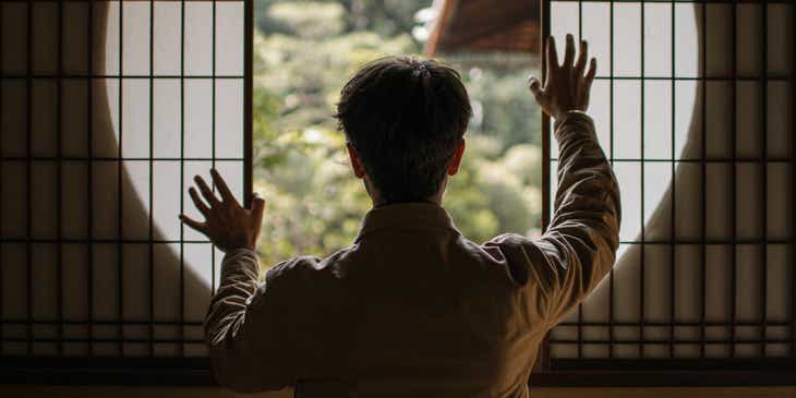 Hombre de espaldas con las manos alzadas frente a una ventana de una habitación zen.