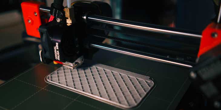 Una máquina de impresión 3D imprimiendo un objeto plateado en un logo para negocios de impresión 3D.