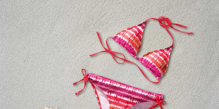Een roze met oranje tie-dye bikini op zand en naast een zeeschelp.