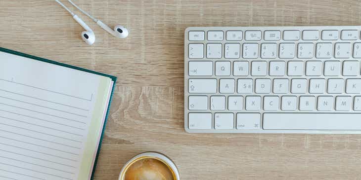 Una tastiera, un quaderno, un caffè macchiato e degli auricolari sulla scrivania di un blogger.