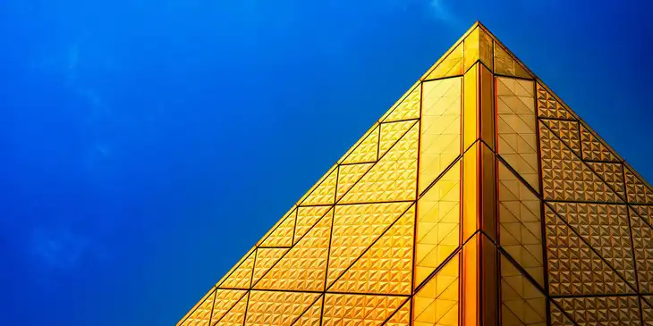 Eine abstrakte, goldene Form, die sowohl an eine Pyramide als auch an ein Hochhaus erinnert, glänzt im Sonnenlicht vor dem blauen Himmel.