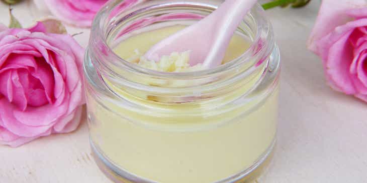 A small jar of body butter with a pink spoon surrounded by pink roses at a body butter business.