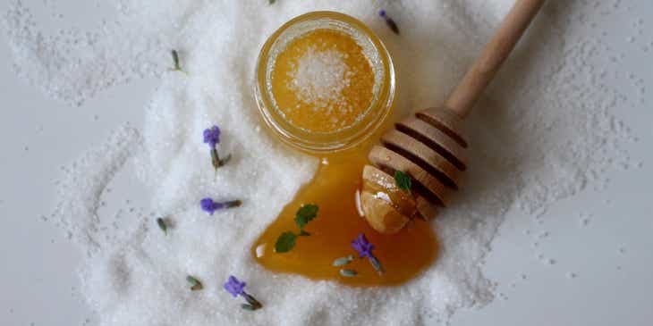Un gommage corporel au miel dans un pot sur du sucre éparpillé et des pétales de lila.