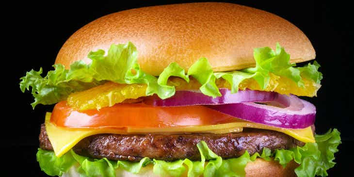 Een heerlijk uitziend broodje hamburger met kaas, tomaat, sla en ui tegen een zwarte achtergrond.
