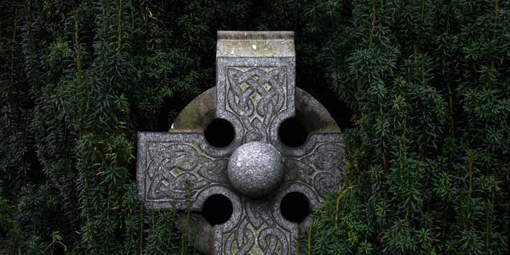 A gray concrete Celtic cross hidden within dark green foliage.