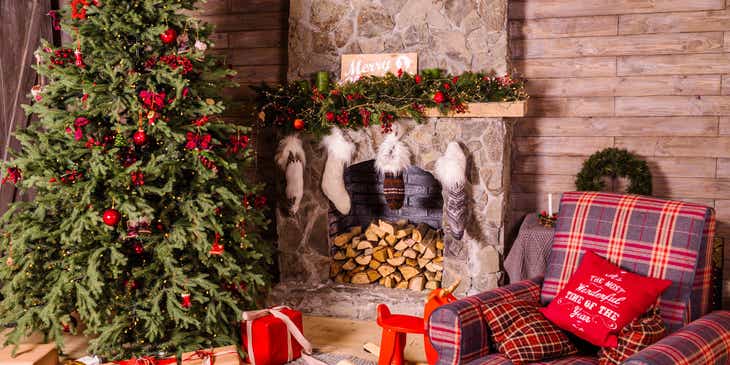 Uma árvore de Natal com presentes embaixo em uma sala com uma lareira com meias penduradas e uma poltrona.