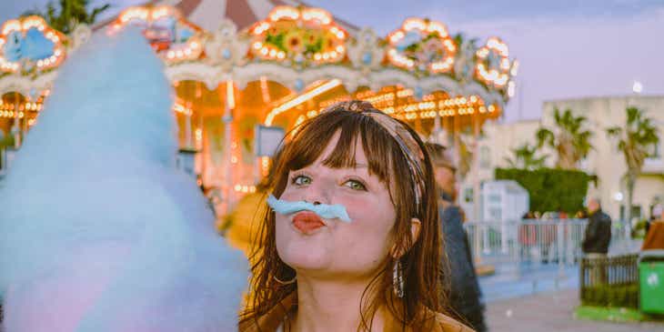 Eine Frau steht neben einem Karussell und hält verspielt ein Stück Zuckerwatte über ihrem Mund, um an einen Schnurrbart denken zu lassen.
