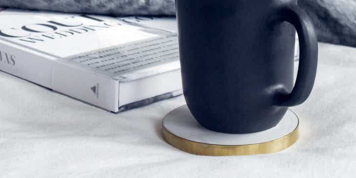 Eine schwarze Tasse steht neben einem Buch und einer Wolldecke auf einem Untersetzer.