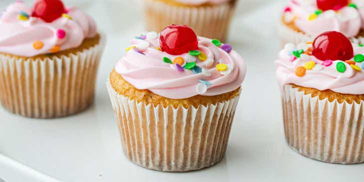 Cupcake pink dengan taburan gula dan ceri merah di atasnya diletakkan di atas piring.