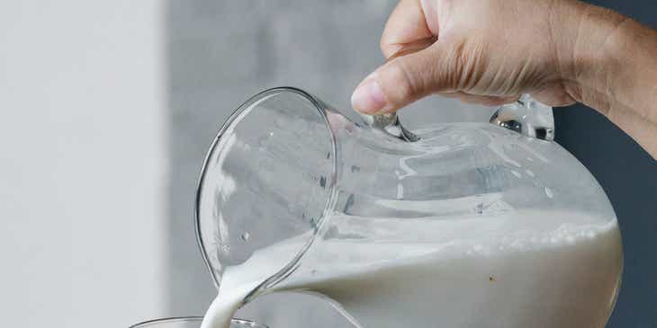 Eine Person gießt frische Milch von einer Molkerei aus einer Kanne in ein Glas.