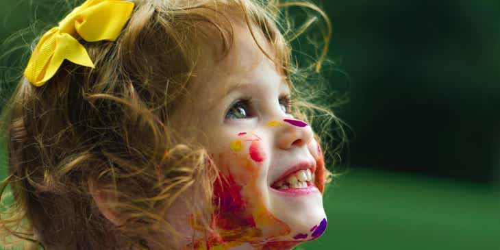 A delighted child covered in paint with a ribbon in her hair.