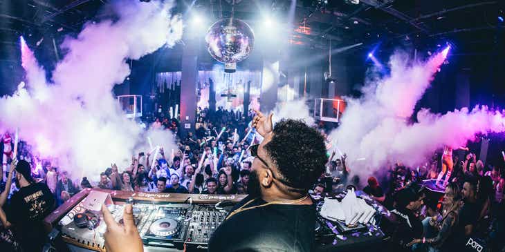 Ein DJ heizt hinter seinem Mischpult in einem Club einem feiernden Publikum ein.