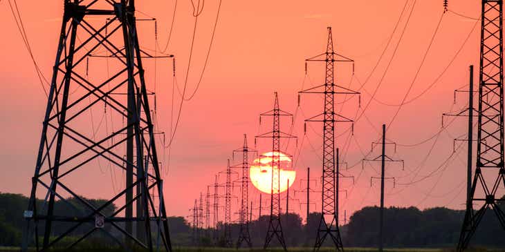 De silhouetten van de hoogspanningsmasten van een elektriciteitsbedrijf tegen een oranje achtergrond met een ondergaande zon.