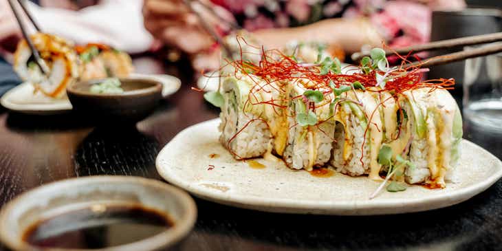 Primo piano di un piatto di sushi con persone sullo sfondo intente a mangiarlo in un ristorante di sushi.