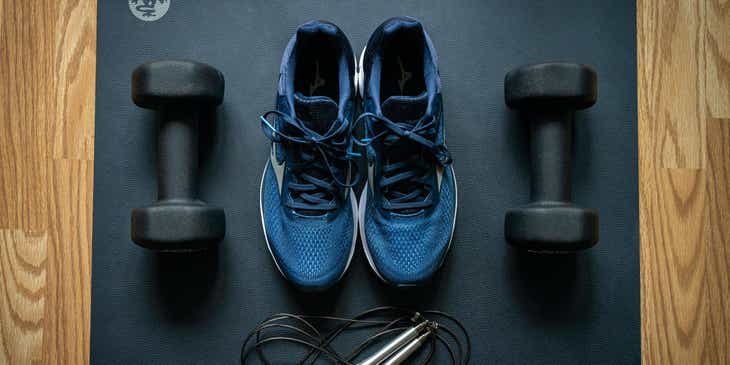 Unas zapatillas deportivas, mancuernas y una cuerda que son parte del equipo de un gimnasio, sobre una esterilla..