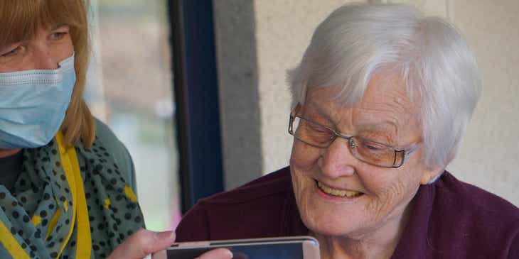 Eine Mitarbeiterin eines häuslichen Pflegediensts zeigt einer älteren, lächelnden Patientin etwas auf ihrem Handy.