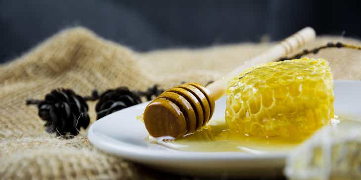 Eine Honigwabe und ein Honiglöffel liegen auf einem weißen Porzellanteller, der auf einer Jutedecke platziert ist.