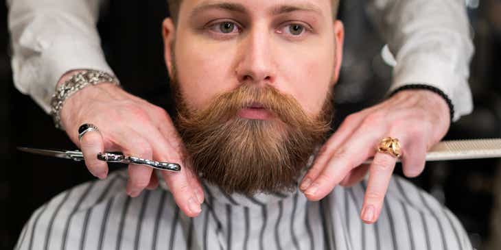 Seorang barber sedang mencukur janggut pelanggan di sebuah barbershop.