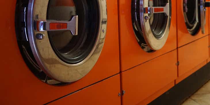 Tiga mesin cuci berwarna oranye di sebuah laundry.