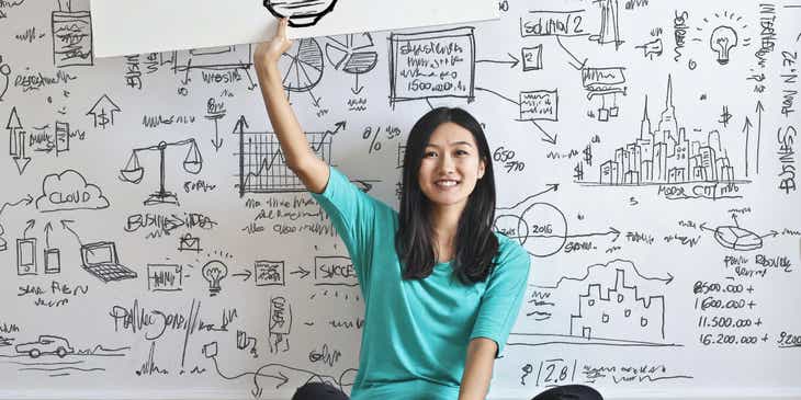 Kobieta siedząca na tle ściany zapisanej innowacyjnymi pomysłami, trzymająca transparent z rysunkiem żarówki.
