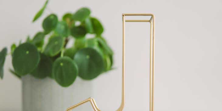 Uma letra "J" feita de fios de latão em cima de uma mesa branca com um vaso de plantas ao lado.