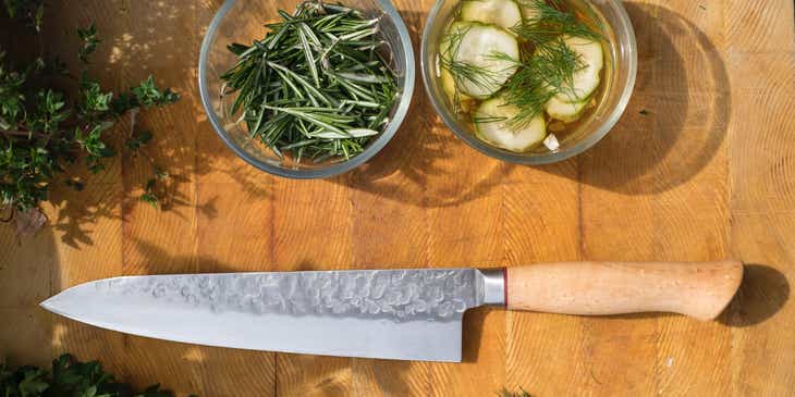 A cooking knife on a wooden table surrounded by chopped and whole herbs and vegetables.
