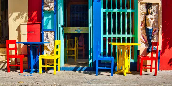 Frente de uma cafeteria colorida em estilo latino com mesas, cadeiras e uma escultura de sereia.