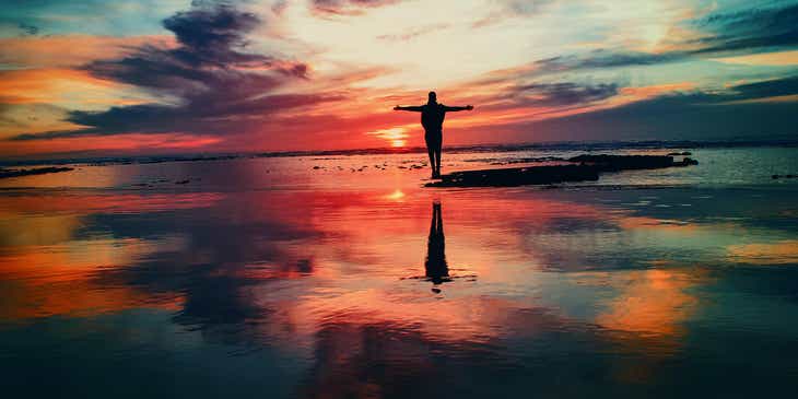 Eine spirituelle Person mit ausgebreiteten Armen steht auf einem Strand und bestaunt den farbenfrohen Sonnenuntergang.