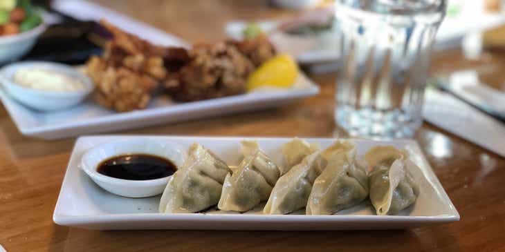 Ein Teller mit fünf Dumplings steht in einem chinesischen Restaurant neben einem Schälchen Soyasoße auf einem Holztisch.