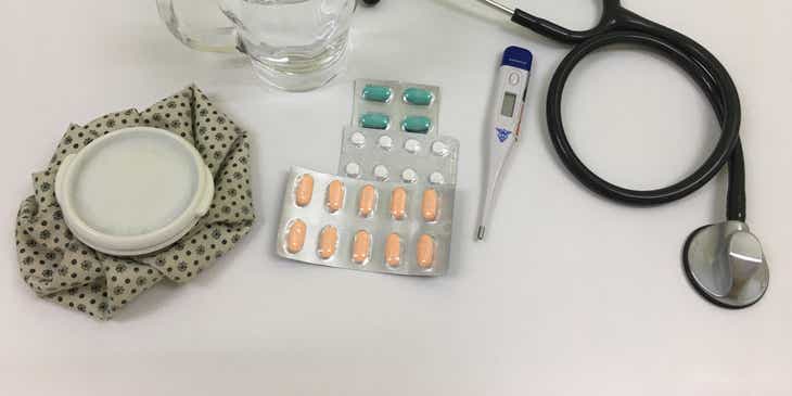 Uno stetoscopio, un termometro, delle pillole, un bicchiere d'acqua e una sacca termica sopra il tavolo di uno studio medico.