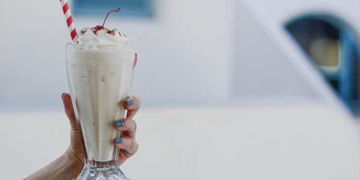 A hand holding a vanilla milkshake in a clear glass.