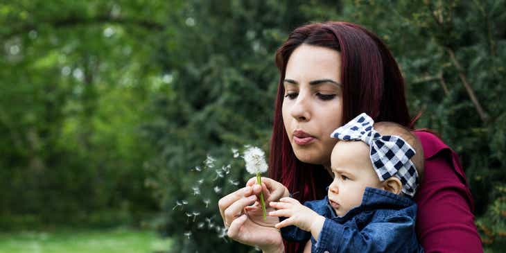 A mom holding her baby daughter while blowing a dandelion.