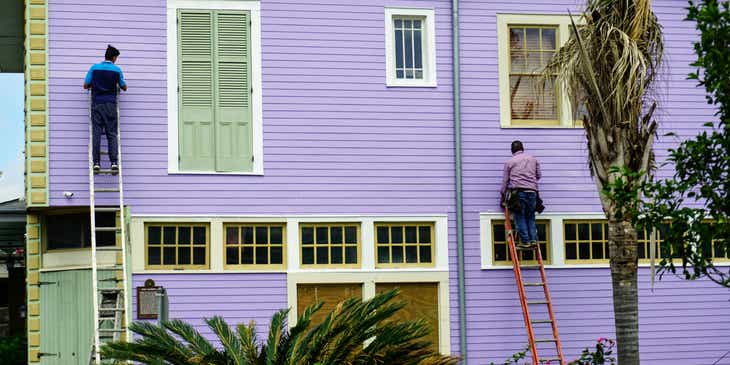 Twee medewerkers van een schildersbedrijf die op ladders een huis paars schilderen.