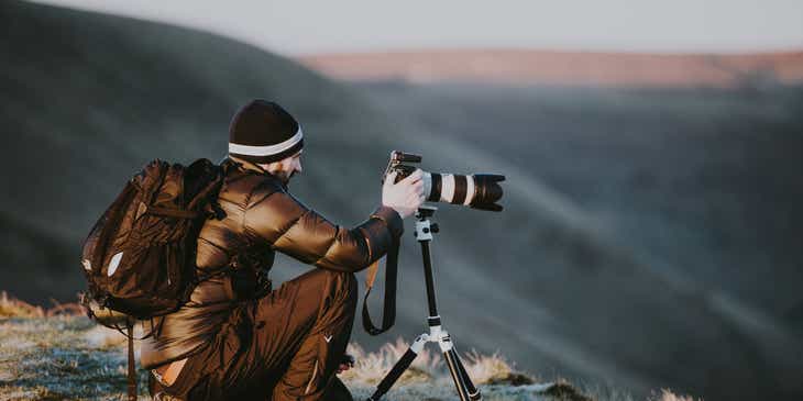 A photographer crouched on mountaintop taking pictures for his photography business.