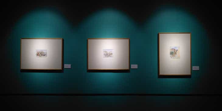Drei goldene Bilderrahmen hängen an der dunkelgrünen Wand einer Kunstgalerie und werden angeleuchtet.