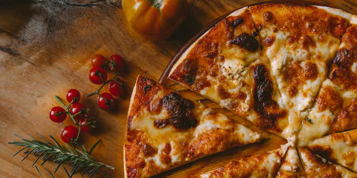 Eine Pizza liegt neben Tomaten und Rosmarin auf einem Holzbrett.